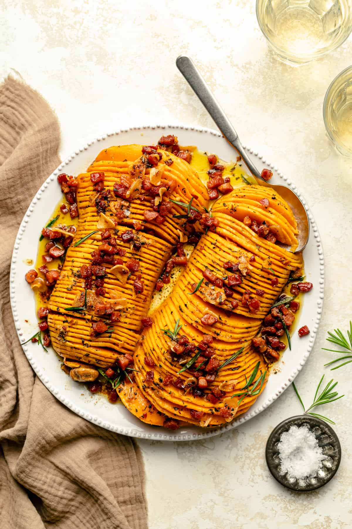 Hasselback butternut squash with bacon and brown butter served on a platter with a spoon and rosemary garnish.
