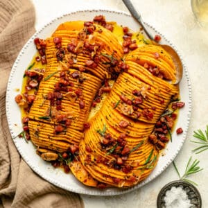 Hasselback butternut squash with bacon brown butter served on a platter with a spoon and rosemary.
