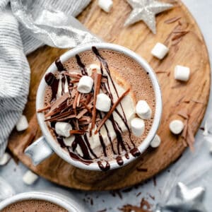 Peppermint hot chocolate in a mug topped with cream, marshmallows, chocolate sauce and served on a wooden plate with a napkin.