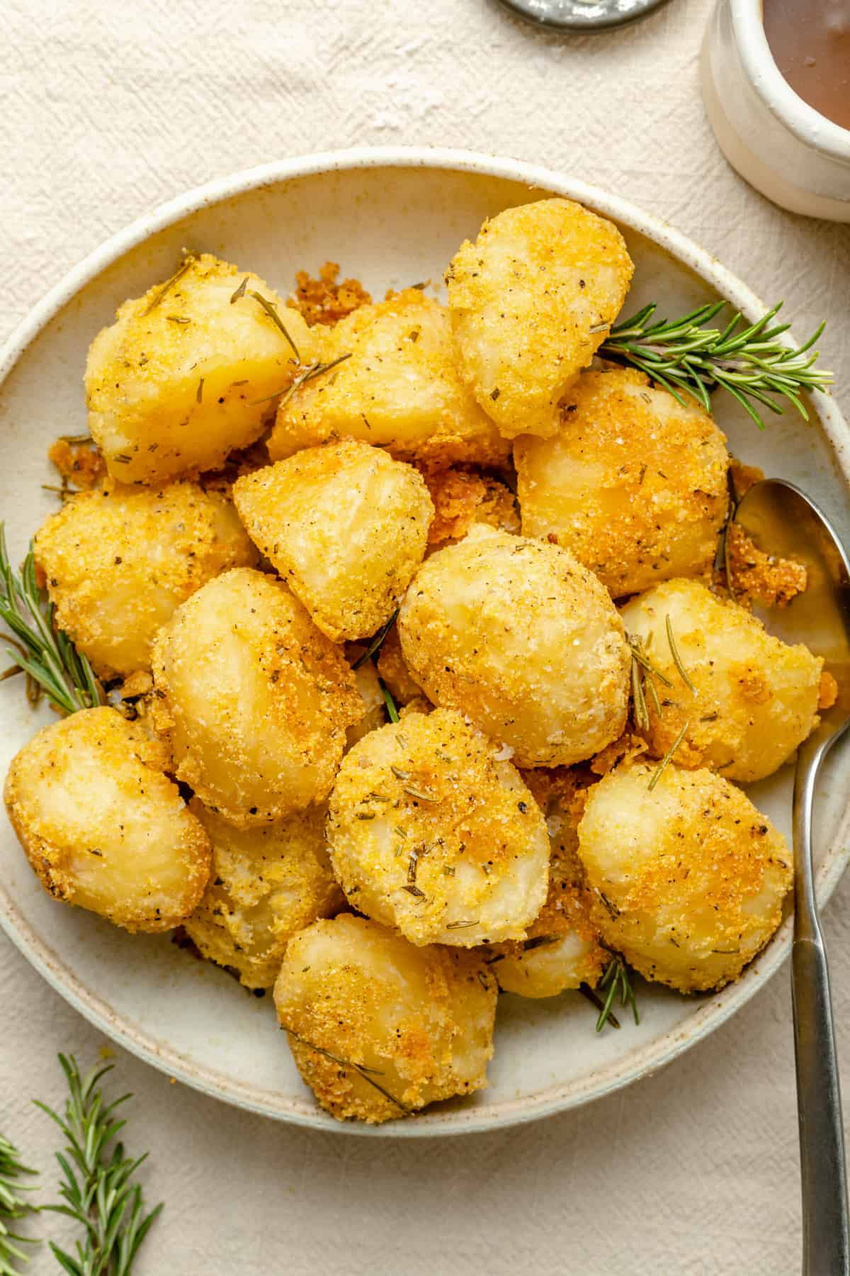 A bowl of crispy rosemary roasted potatoes with rosemary sprigs and a serving spoon with a jug of gravy to the side.