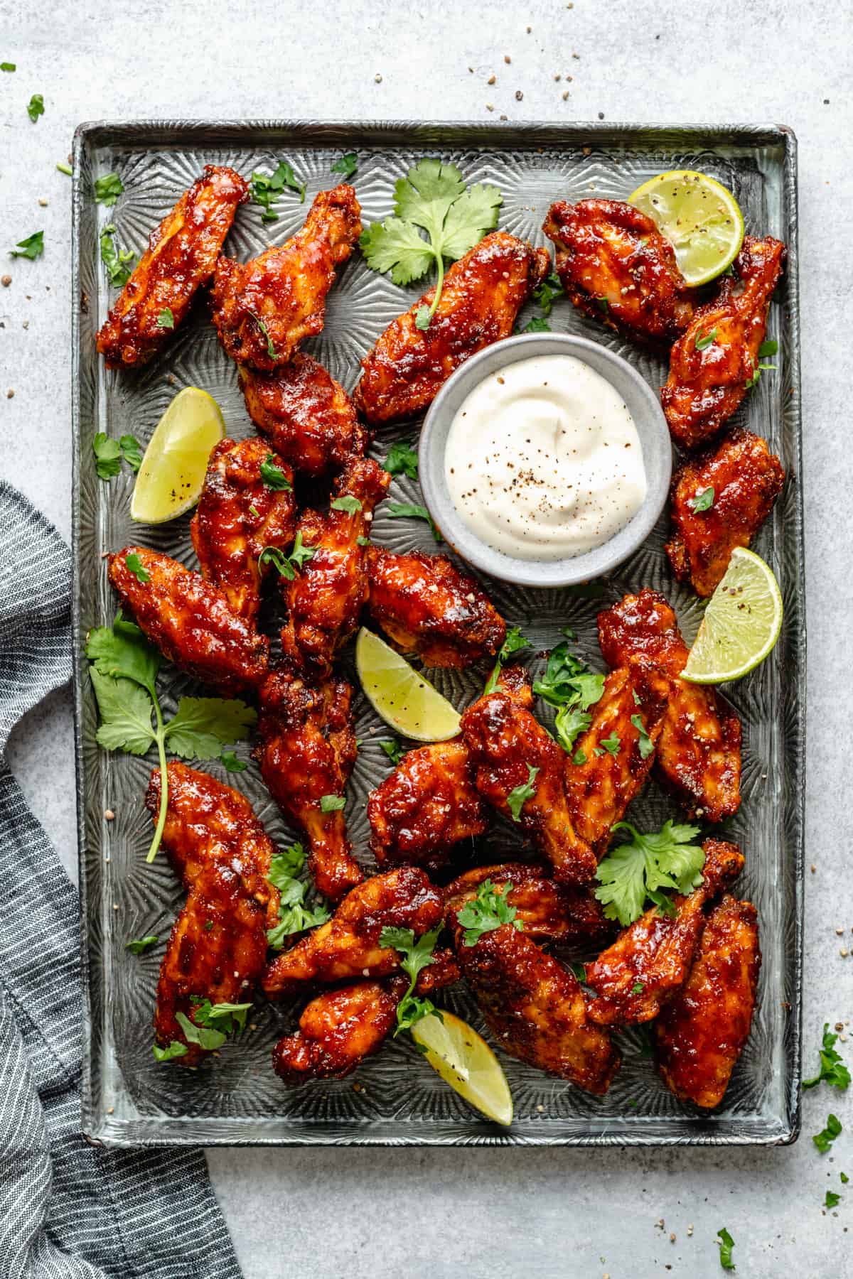 An array of sticky spicy baked chicken wings on a metal tray with lime wedges and coriander leaves served with a small bowl of dip.