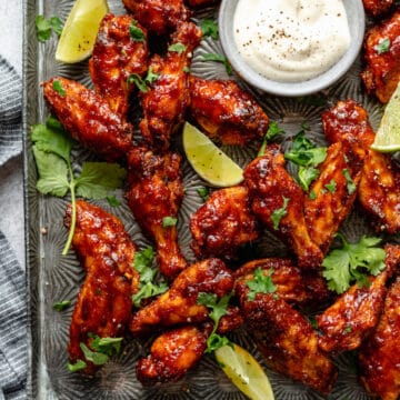 Sticky spicy baked chicken wings on a metal tray with lime wedges with a creamy dipping sauce in a small bowl.