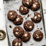 An array of chocolate chip mint cookies on a parchment lined tray with peppermint bark shards around.