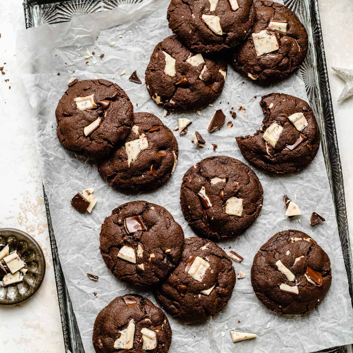 An array of double chocolate chip mint cookies on a baking sheet with broken peppermint bark around.