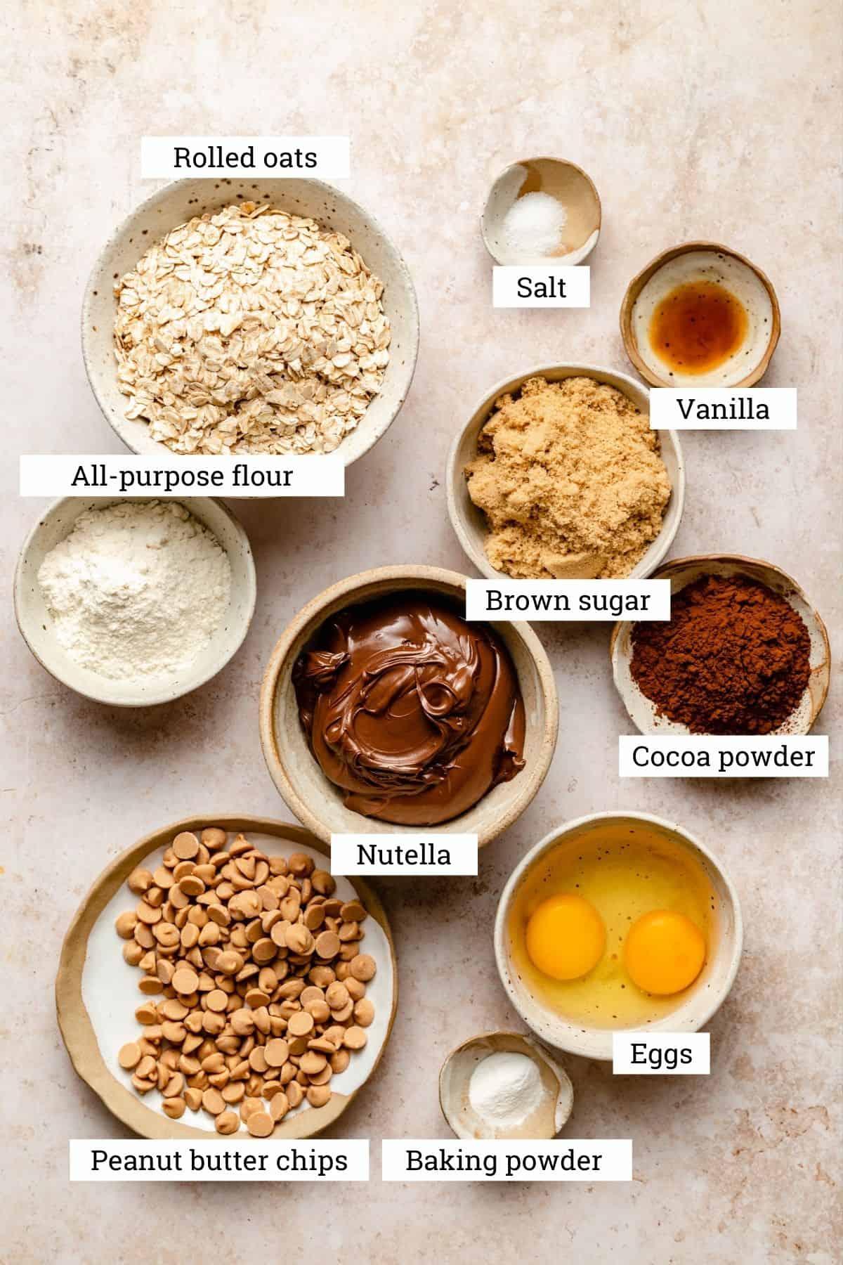 The ingredients for the cookies in various bowls including nutella, oats and peanut butter chips.