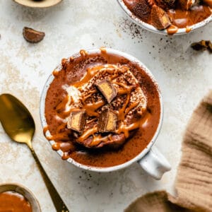 Peanut butter hot chocolate topped with whipped cream, peanut butter and peanut butter cups with a spoon to the side.