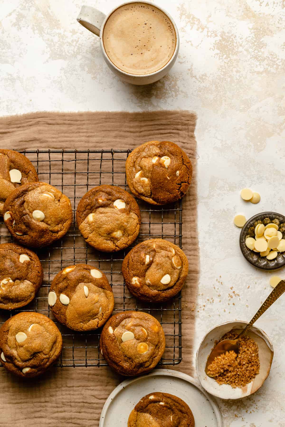 An array of white chocolate gingerbread swirl cookies on a cooling rack with a coffee, chocolate buttons and sugar to the side.