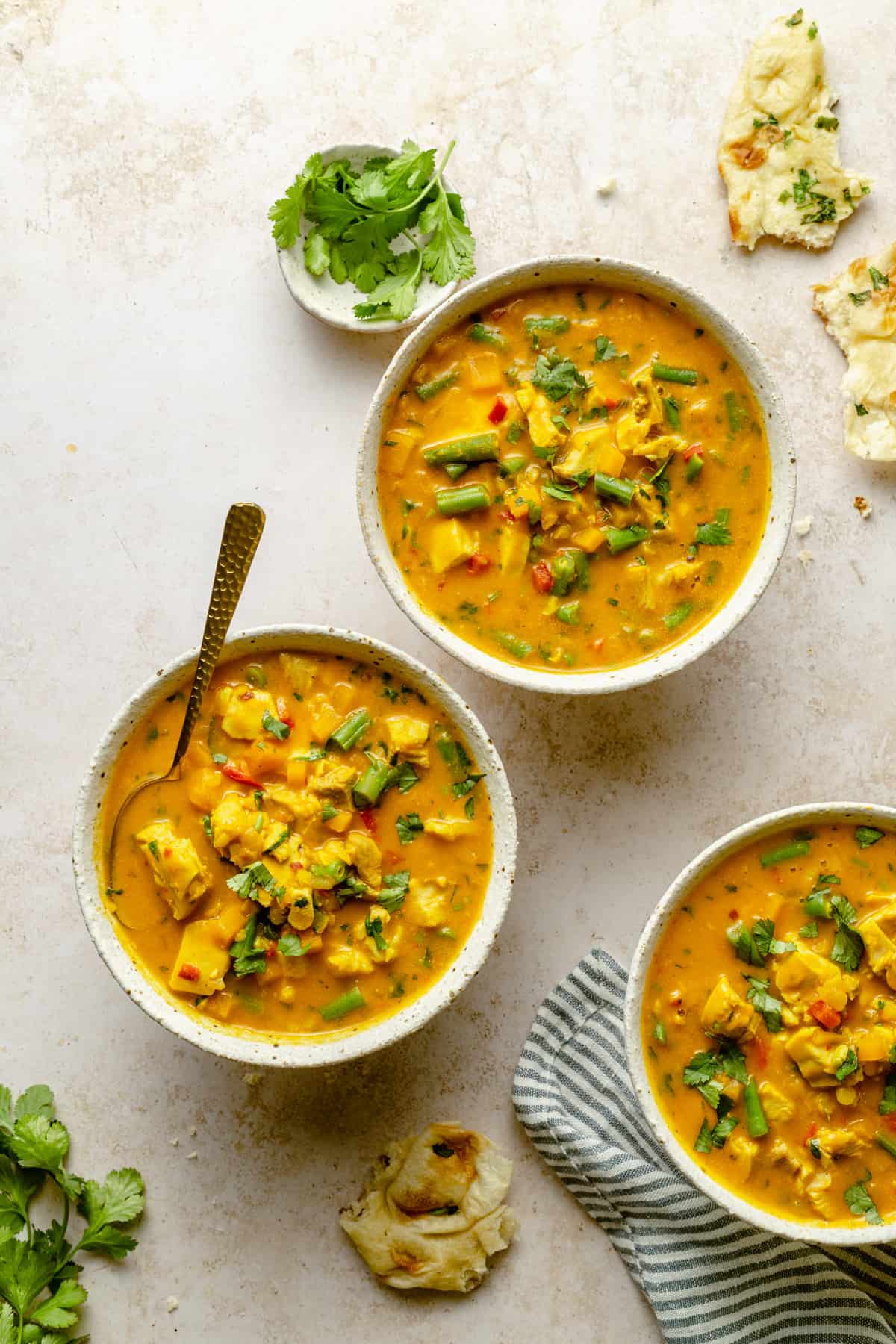 Three bowls of creamy coconut curry chicken soup with a spoon, naan, cilantro and napkin.