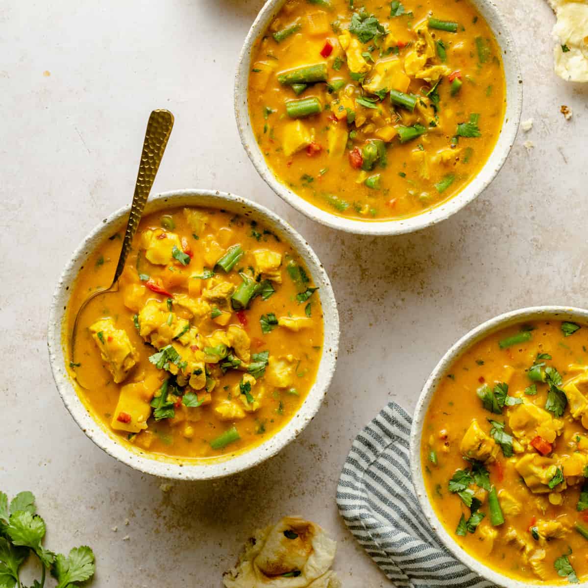 bowls of coconut curry chicken soup with a spoon, napkin, naan and cilantro around by.