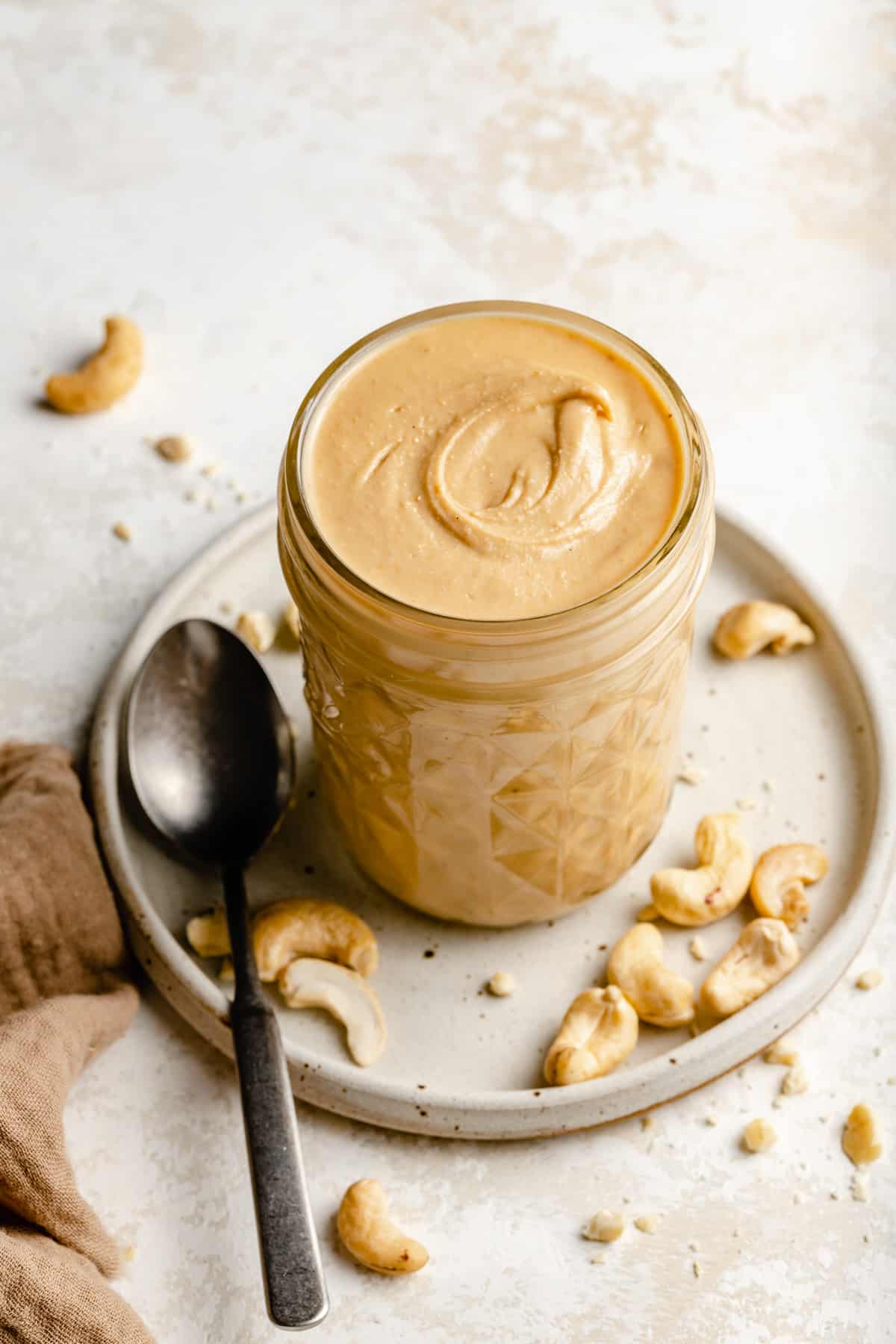 Cashew butter in a jar with a swirl on the top, sitting on a plate with raw cashew nuts and a spoon to the side.