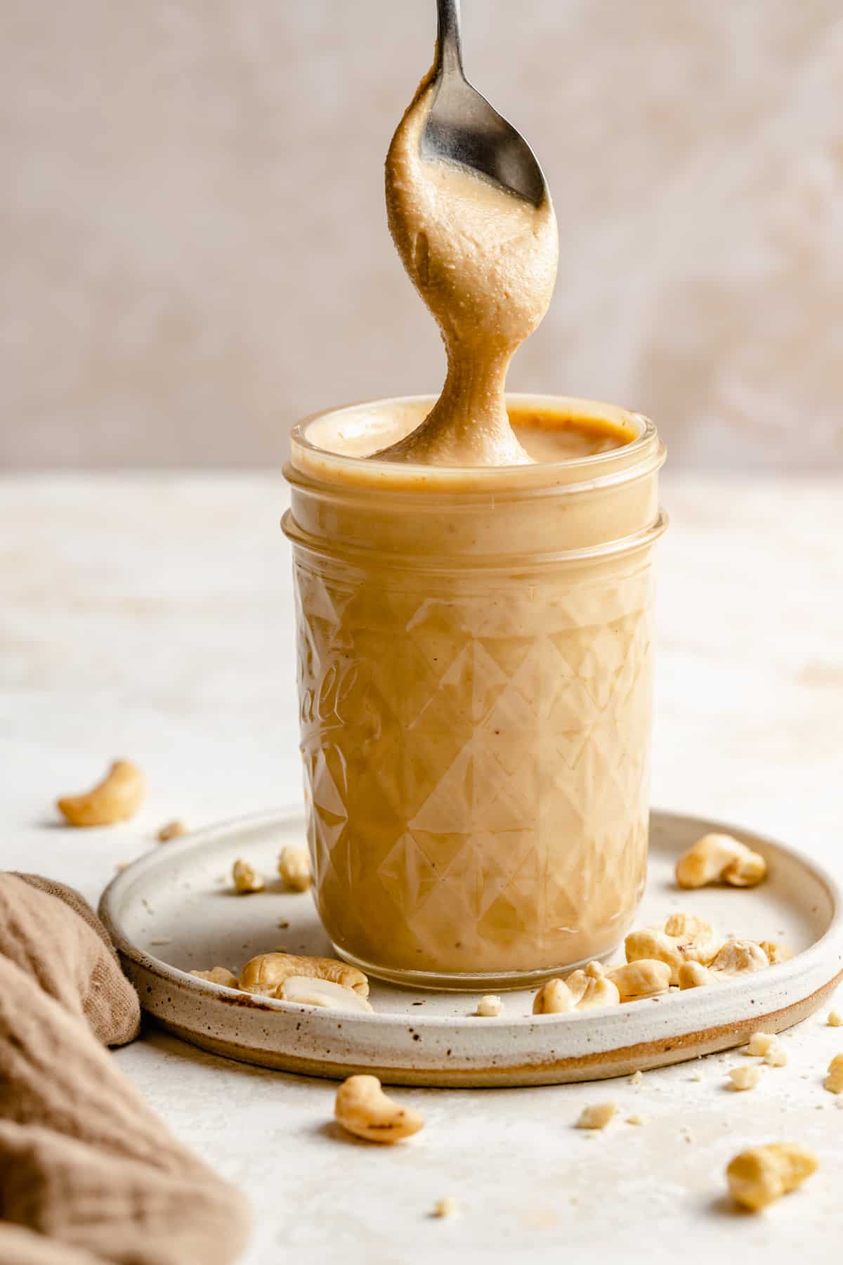 Cashew butter in a jar on a plate with raw cashews and a spoon lifting up a big drip.
