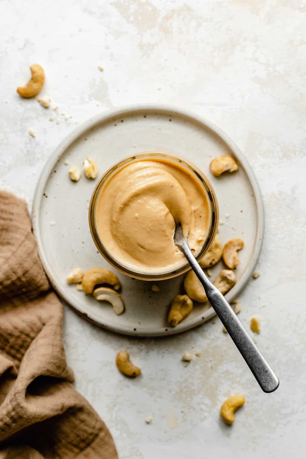 a spoon laying across the top of a jar of cashew butter with raw cashews scattered around.
