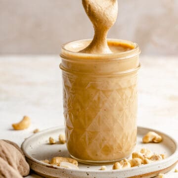 Cashew butter in a jar on a plate with raw cashews around it and a spoon lifting the cashew butter out of the jar in a big drip.