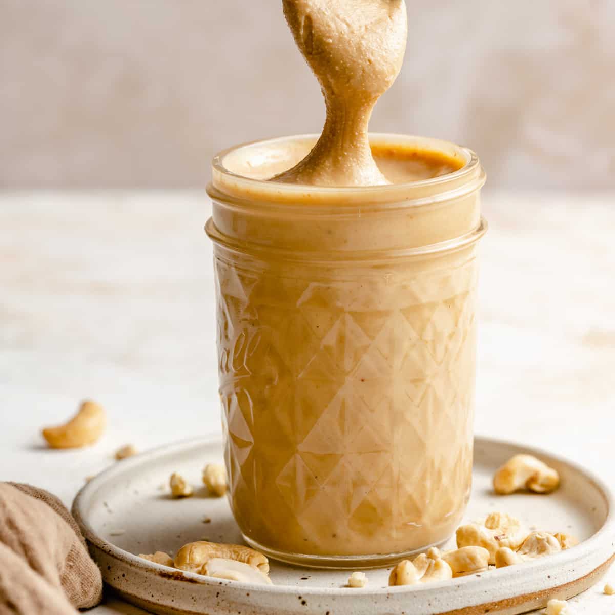 A jar of cashew butter with a spoon pulling a drizzle up, surrounded in roasted cashew nuts.