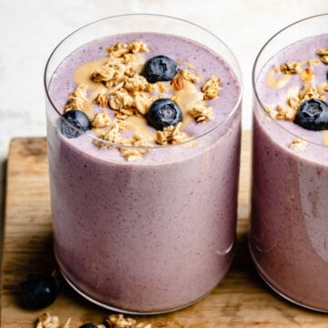 strawberry blueberry smoothie in two glasses on a wooden board with granola, cashew butter and blueberries on top.
