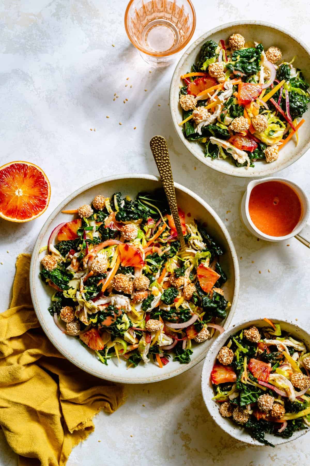 Three bowls of kale citrus salad with a jug of blood orange dressing and glasses of water and a gold fork. 