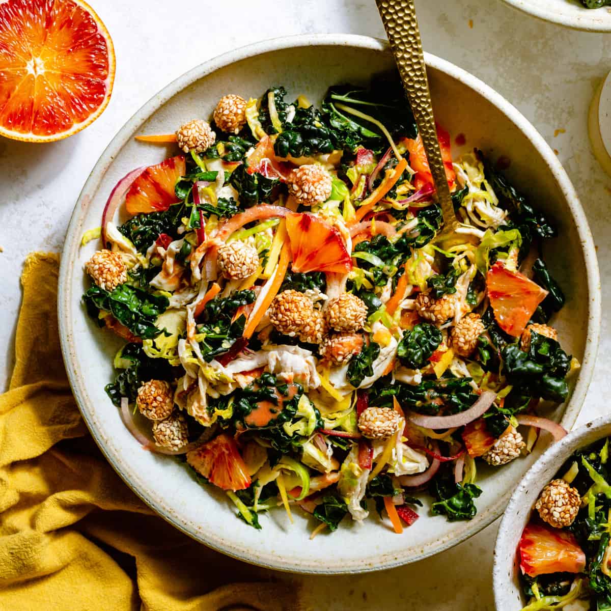 A bowl of kale chickpea salad with chicken, a fork napkin and blood orange to the side. 