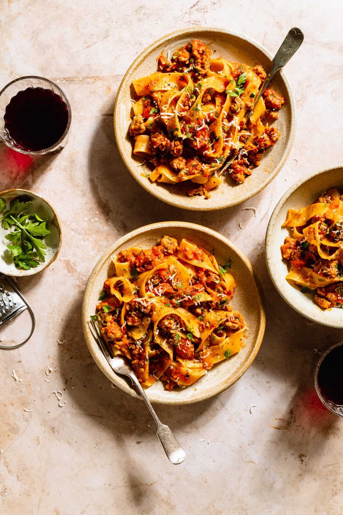 Dinner setting of three bowls of sausage ragu with forks and red wine in glasses with a dish of parsley on the side.