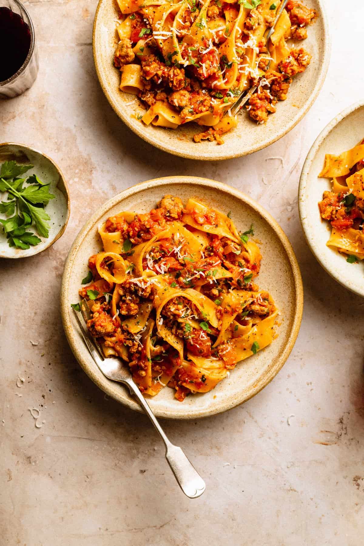 Sausage ragu with pappardelle in bowls, a fork on the side of a bowl and small dish with herbs with a glass of red wine.