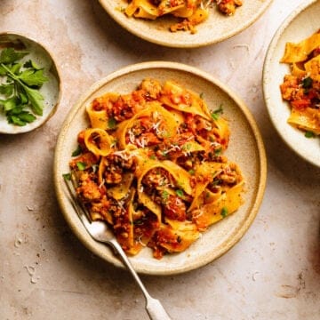 Sausage ragu with pappardelle in bowls with a fork and herbs in a bowl nearby.