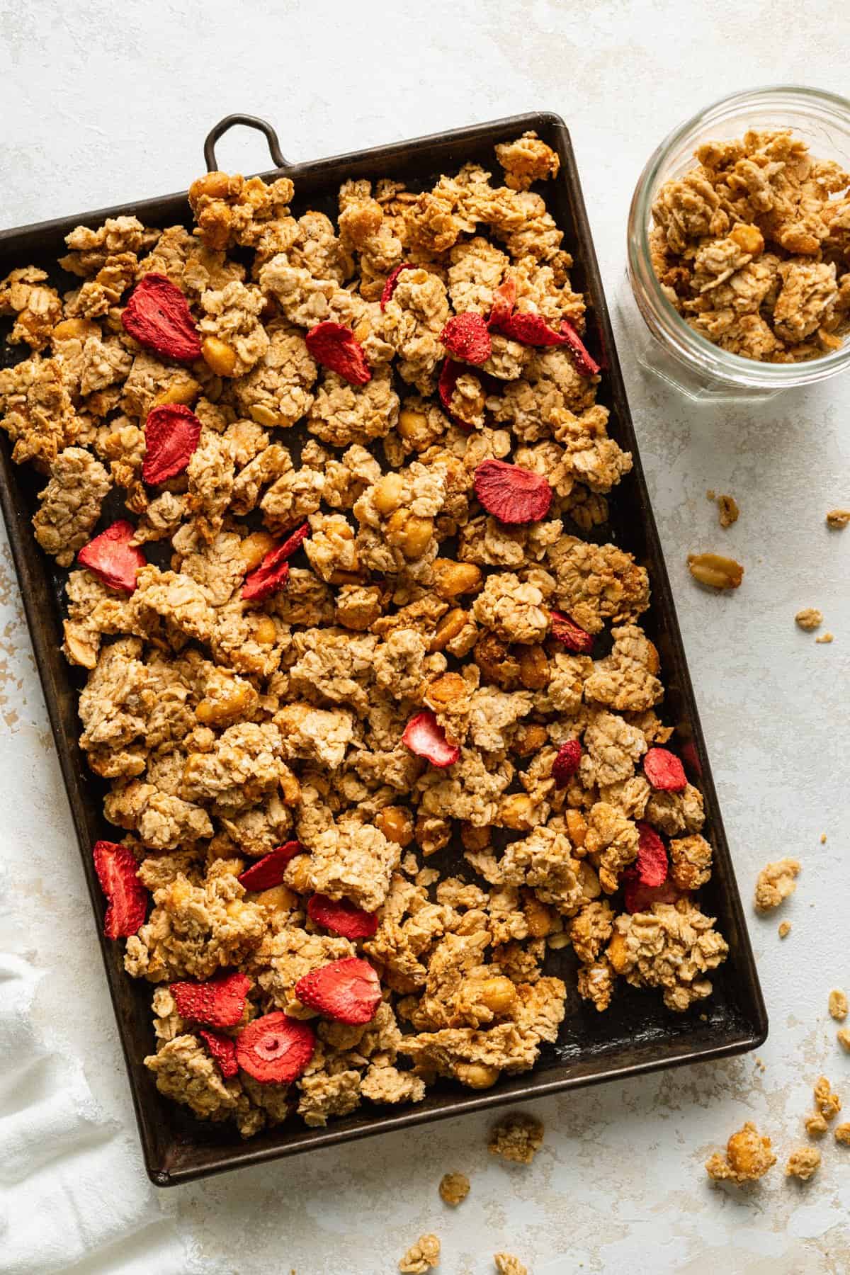 Peanut butter granola clusters with freeze dried strawberries on a baking sheet with a jar of it to the side.