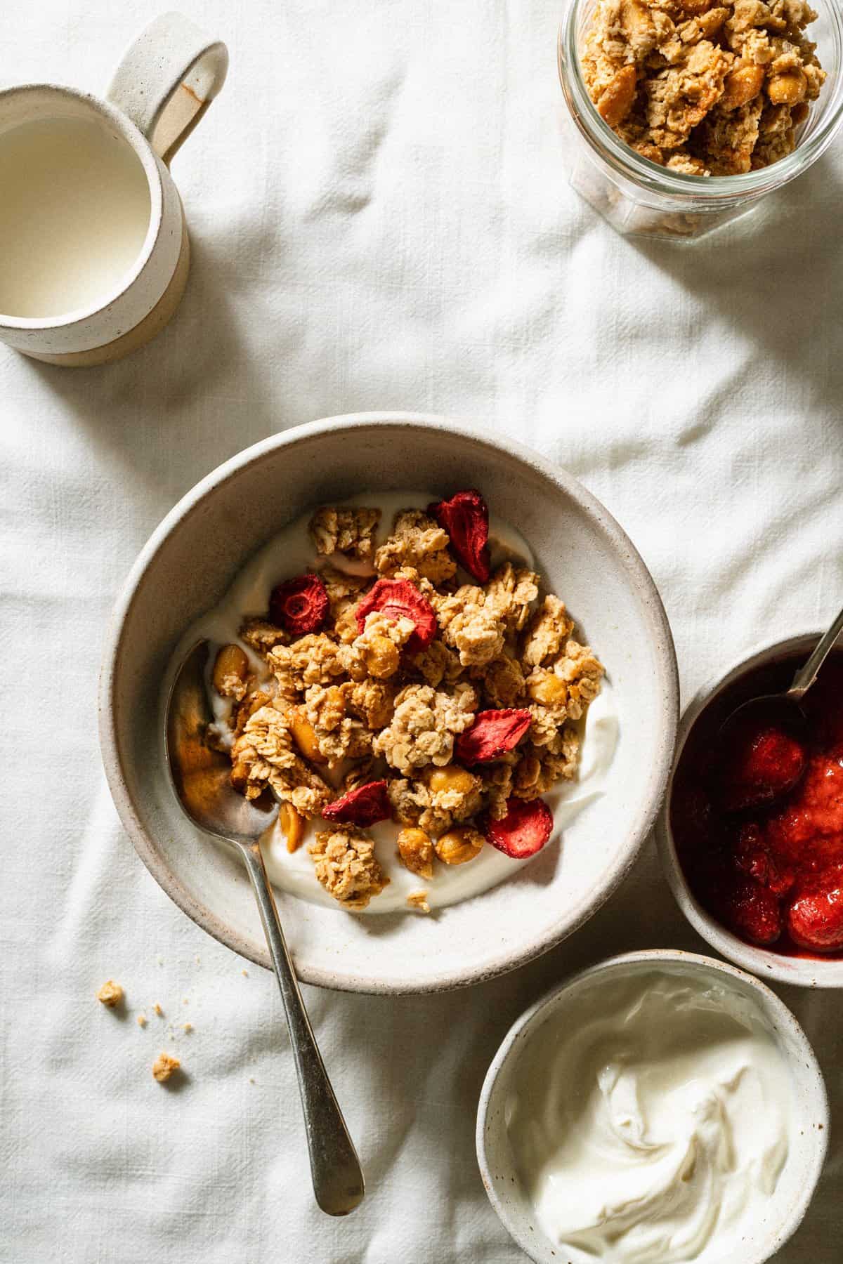 A morning breakfast scene with a bowl of granola served on yoghurt with a spoon and some compote in a dish to the side.