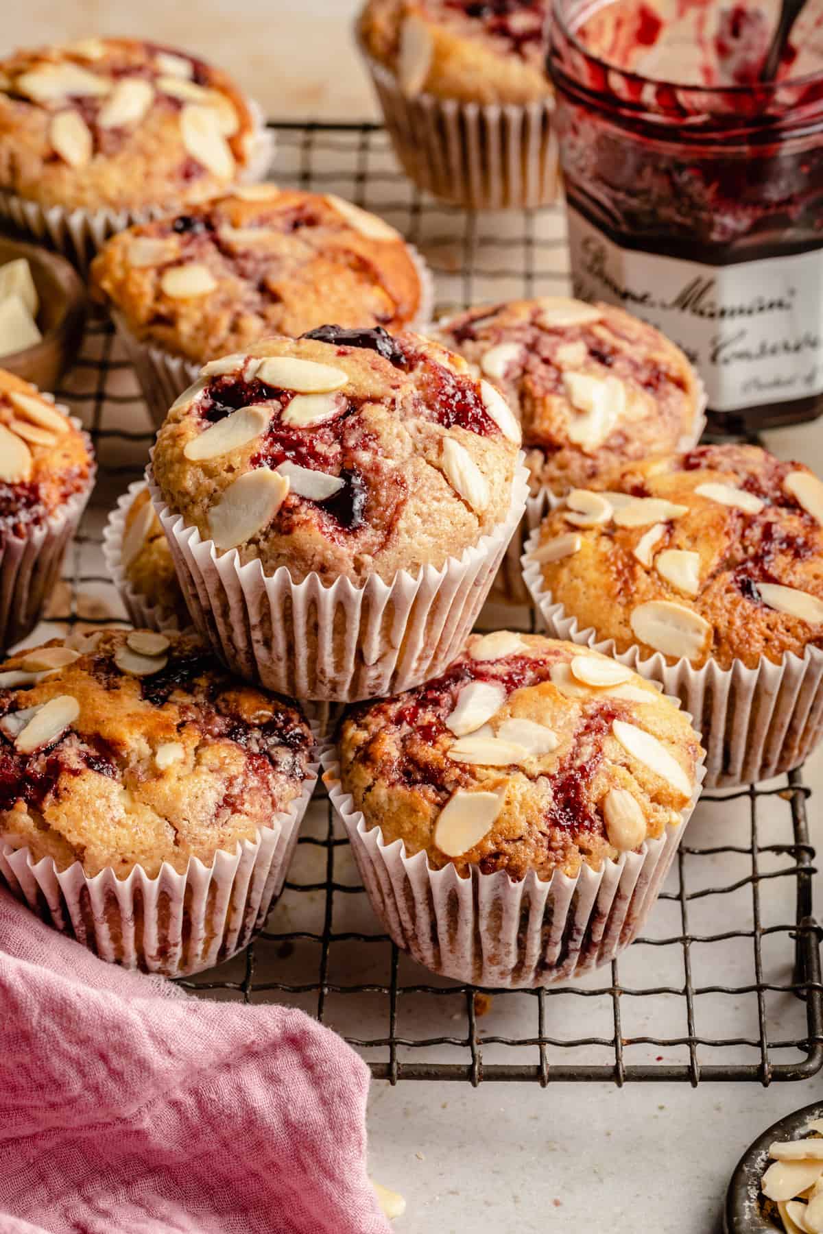 An array of cherry sourdough muffins on a wire rack stacked up with a jar of jam nearby.