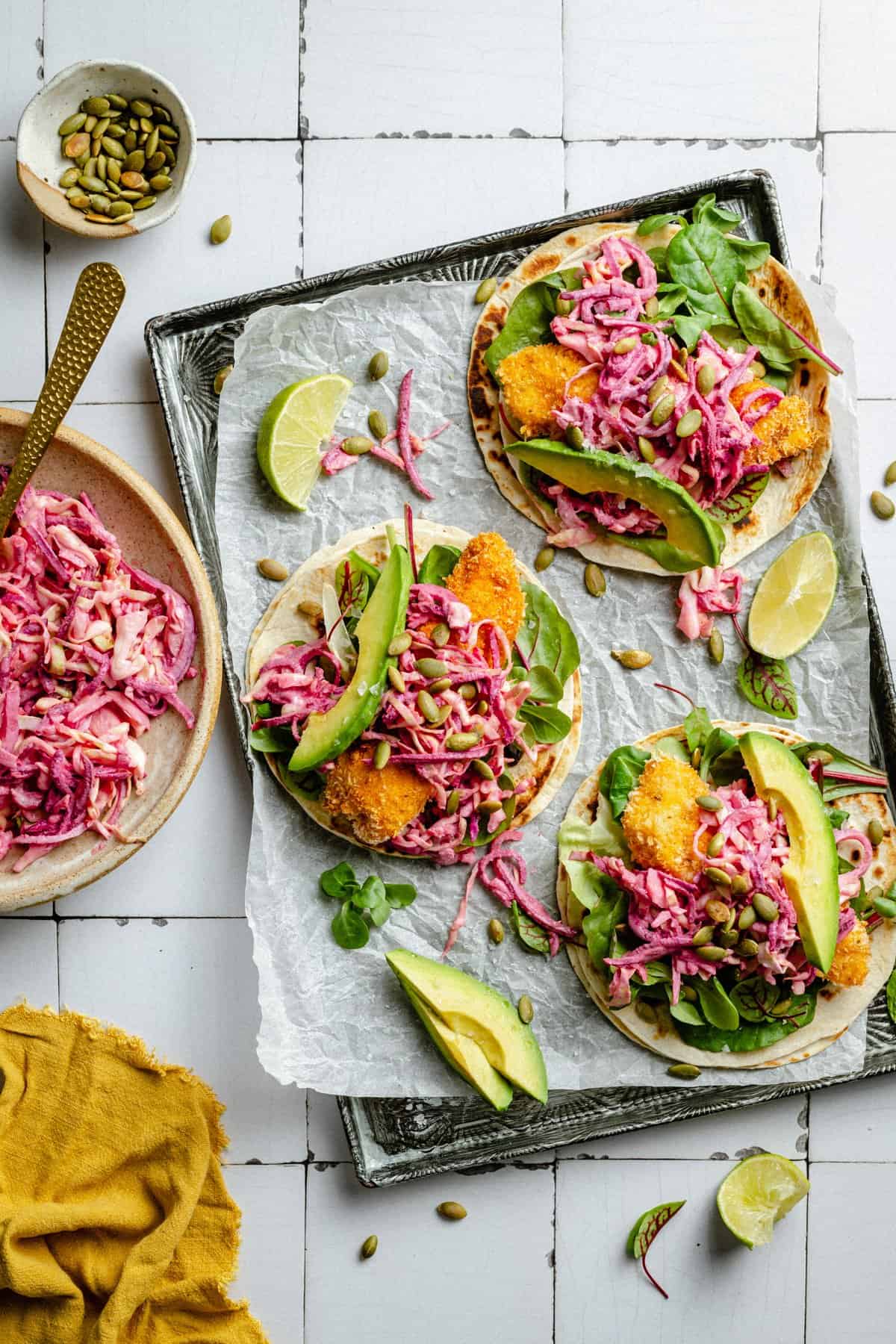 A tray lined with parchment paper with three assembled crispy chicken tacos and a bowl of slaw.