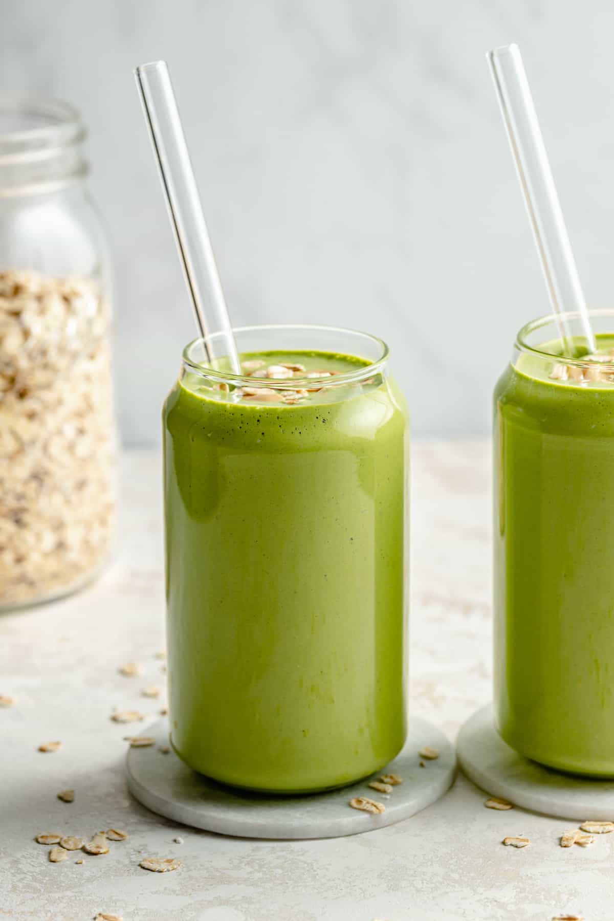 Two glasses of banana spinach smoothie with glass straws with a jar of oats.