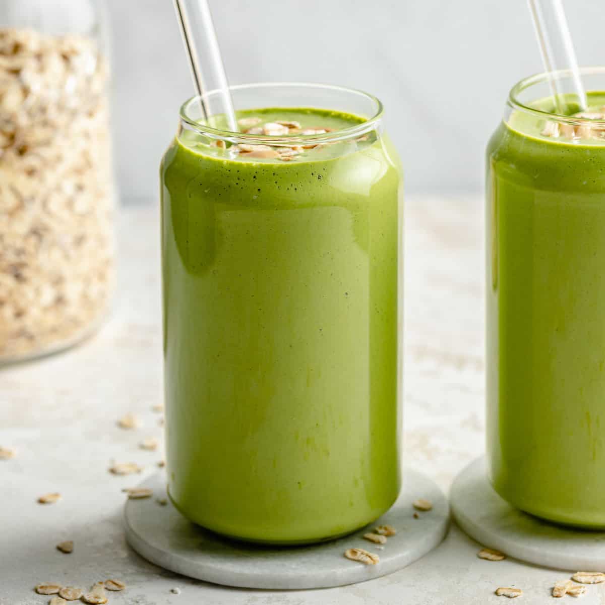 Two glasses of banana spinach smoothie on marble coasters with straws and oats on top.