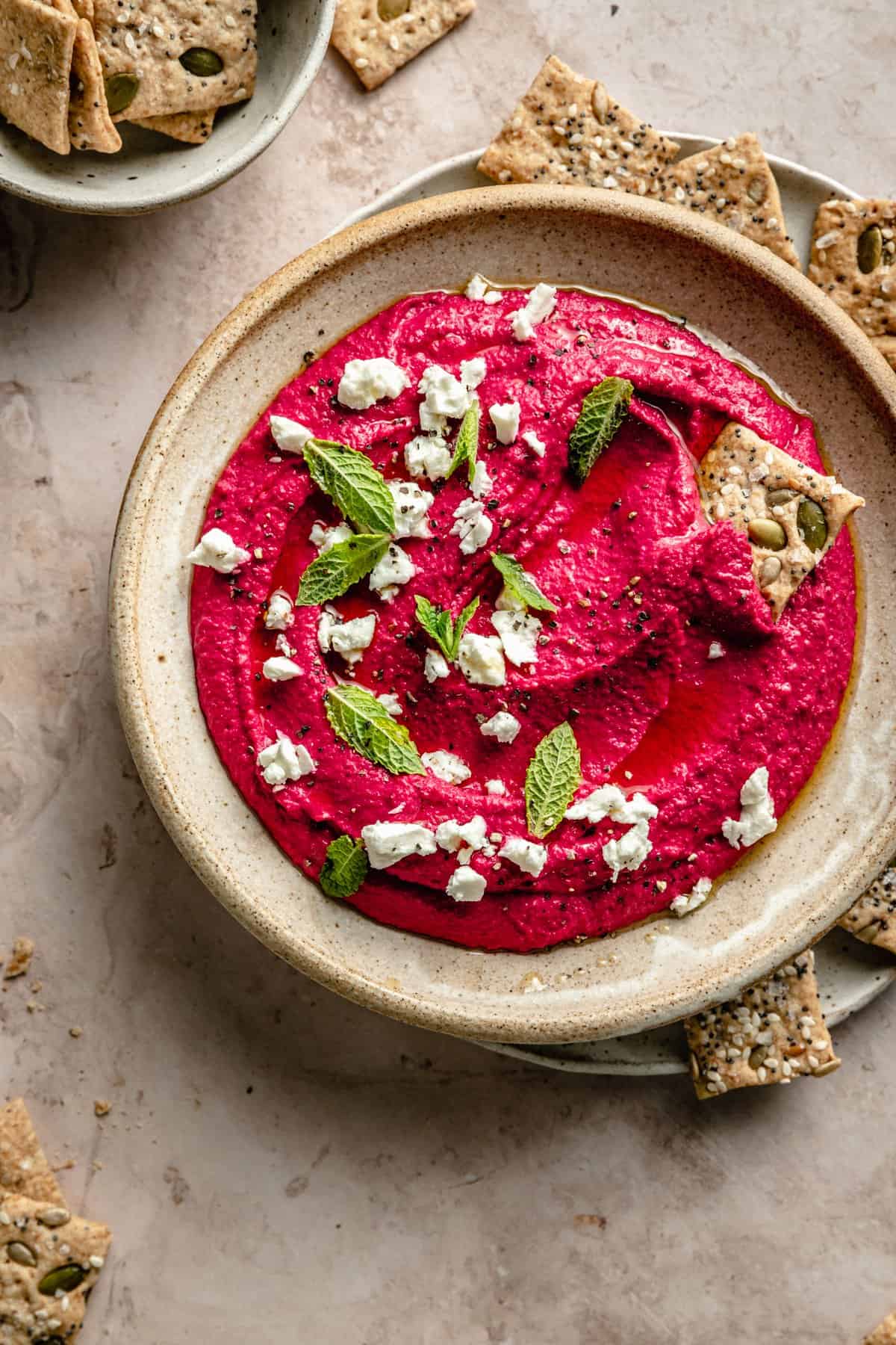 A bowl of beetroot hummus on a plate surrounded with crackers.