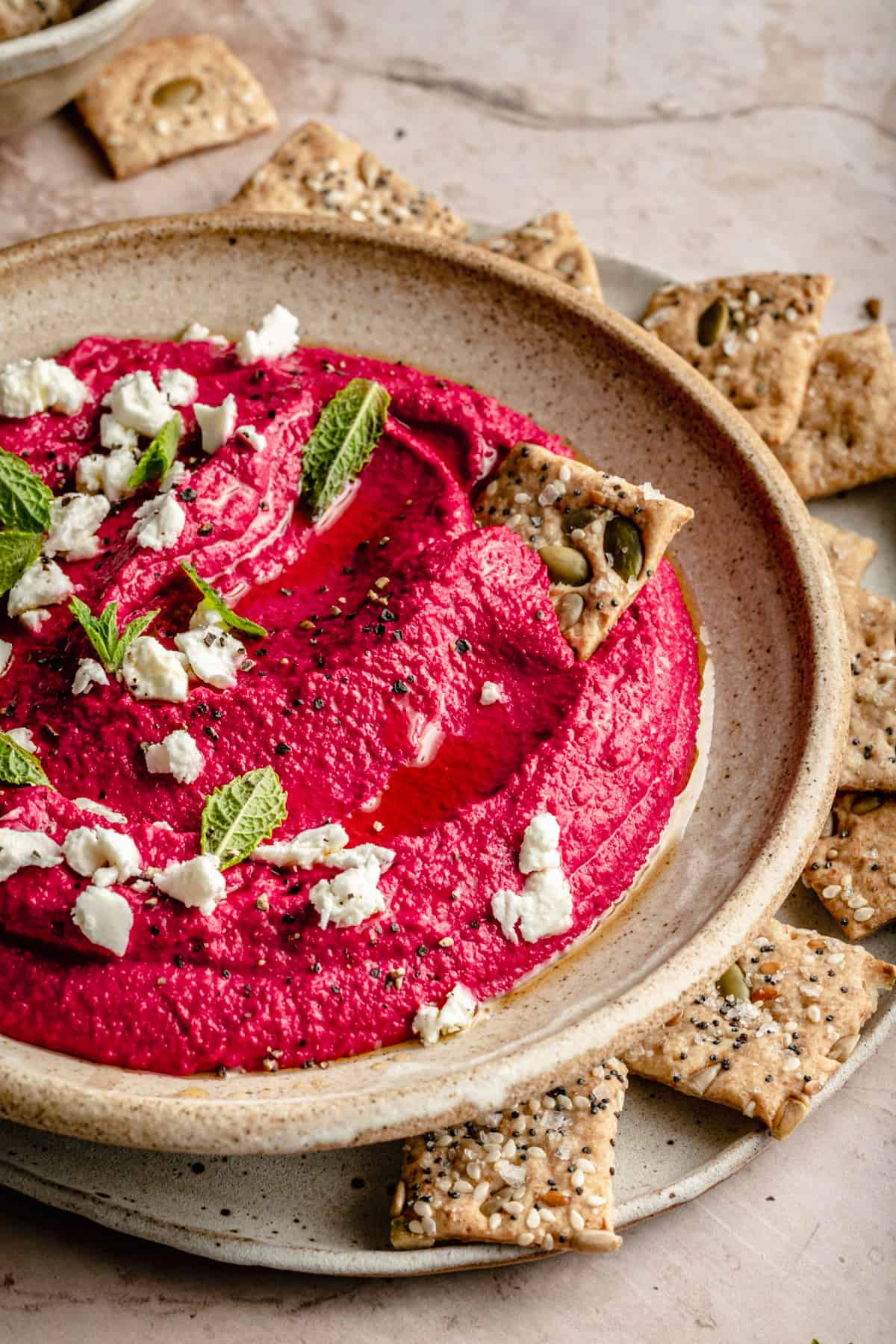 A bowl of beetroot hummus with feta mint leaves and a cracker dipping in it.