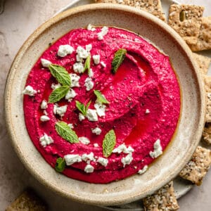 Beetroot hummus in a bowl with feta, mint and olive oil on top with crackers around.