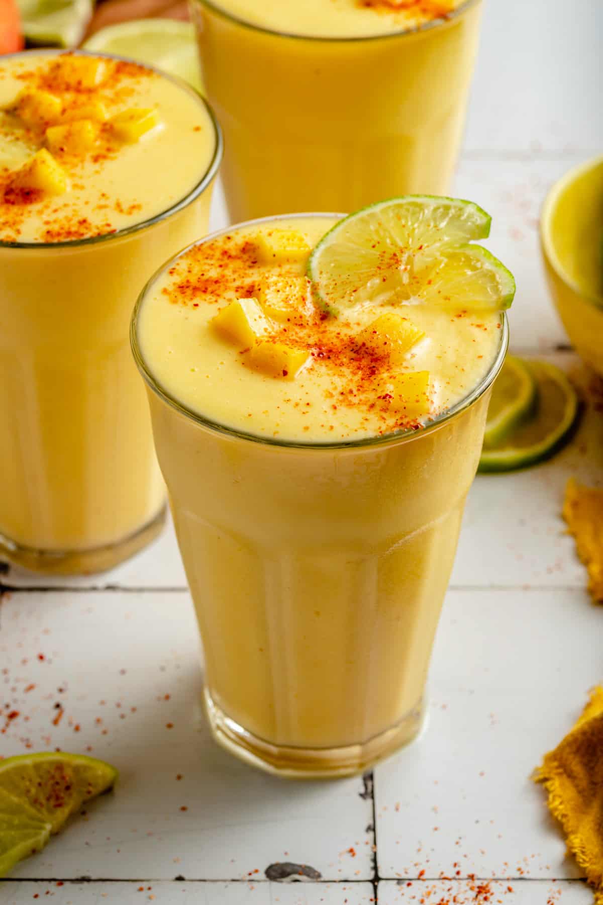 Glasses of mango pineapple smoothie on a tiled surface with lime and mango around.