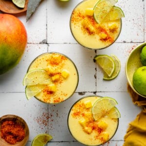 Mango pineapple smoothie in three glasses with mango, lime and tajin on top.