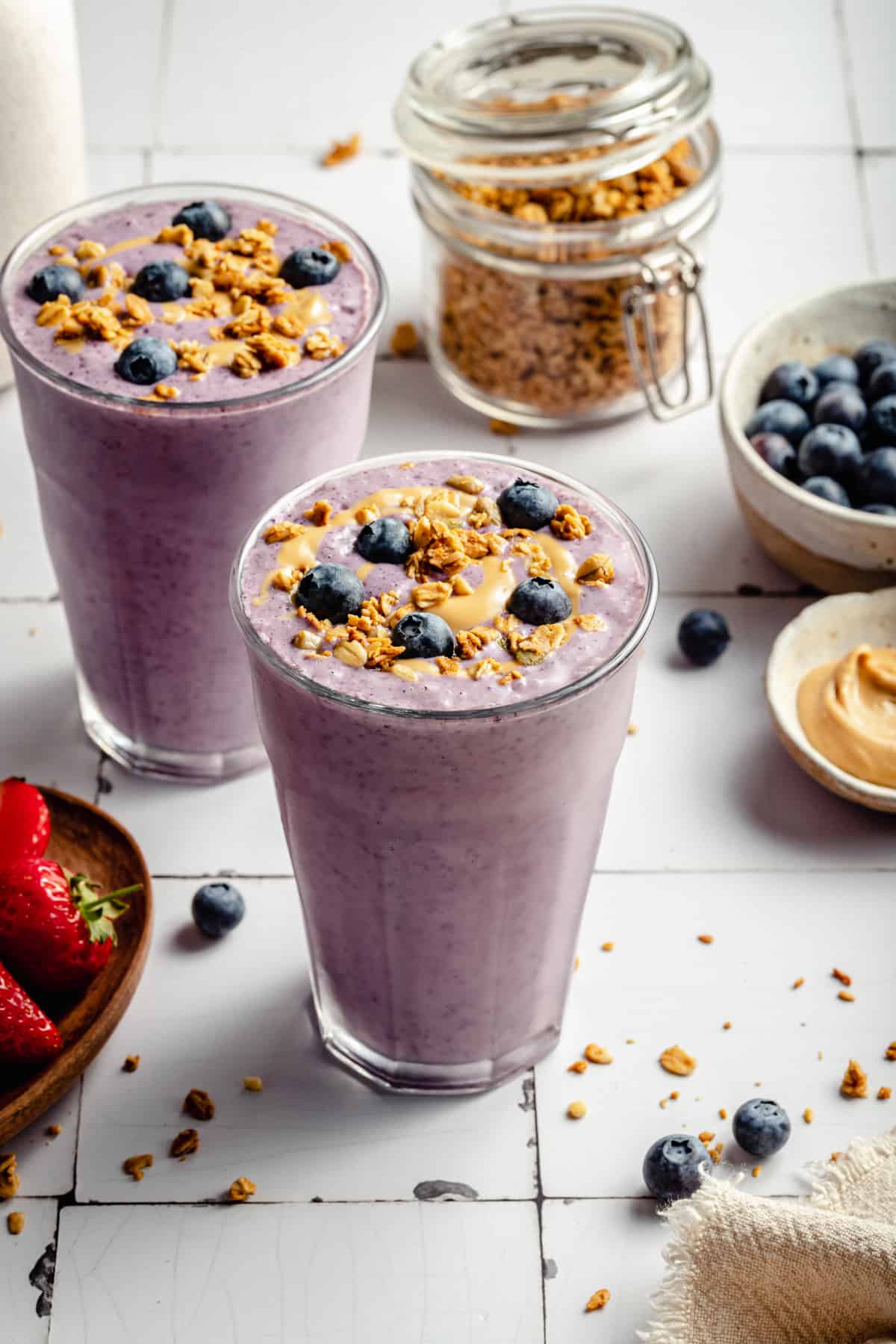 Strawberry blueberry smoothie in two glasses with granola, cashew butter and blueberries around.