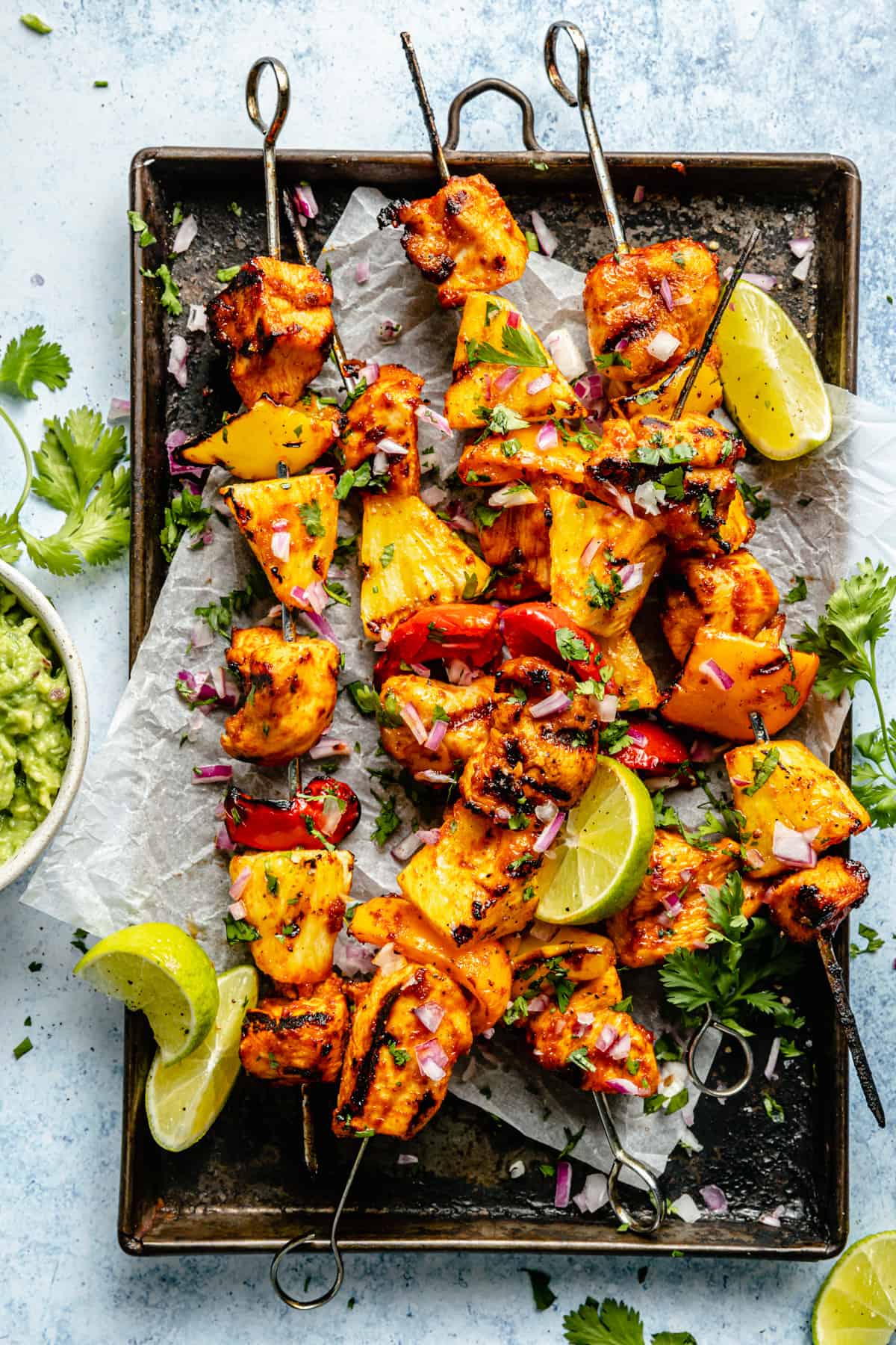 Pineapple chicken kabobs on a baking sheet with guacamole and limes around.