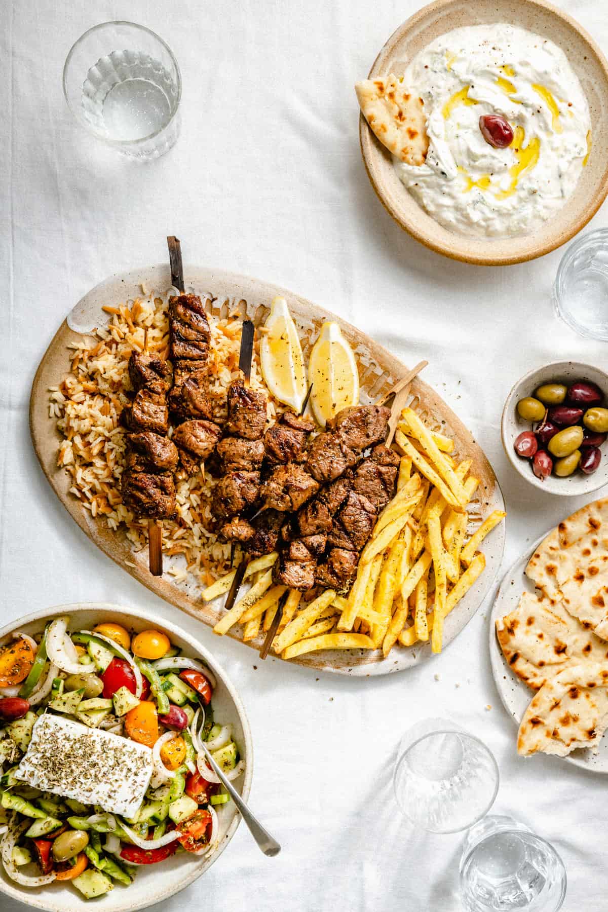 A spread of lamb souvlaki surrounded with tzatziki, olives and greek salad.