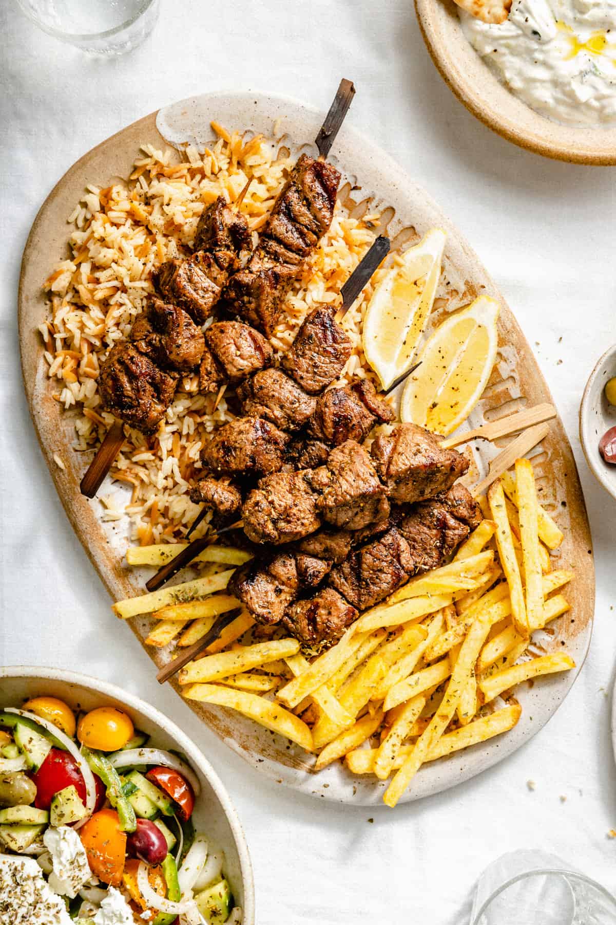 Lamb souvlaki skewers served atop orzo rice and fries on a platter.