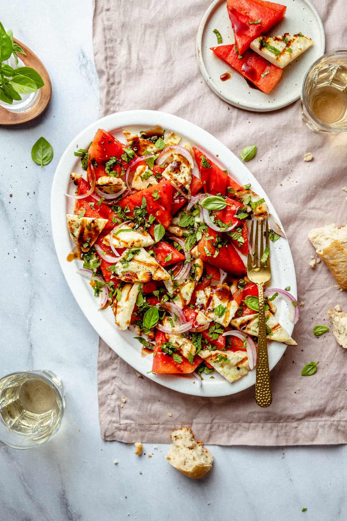 Watermelon basil salad on a platter with grilled halloumi and crusty bread broken up around the platter. Served with glasses of wine on a napkin.