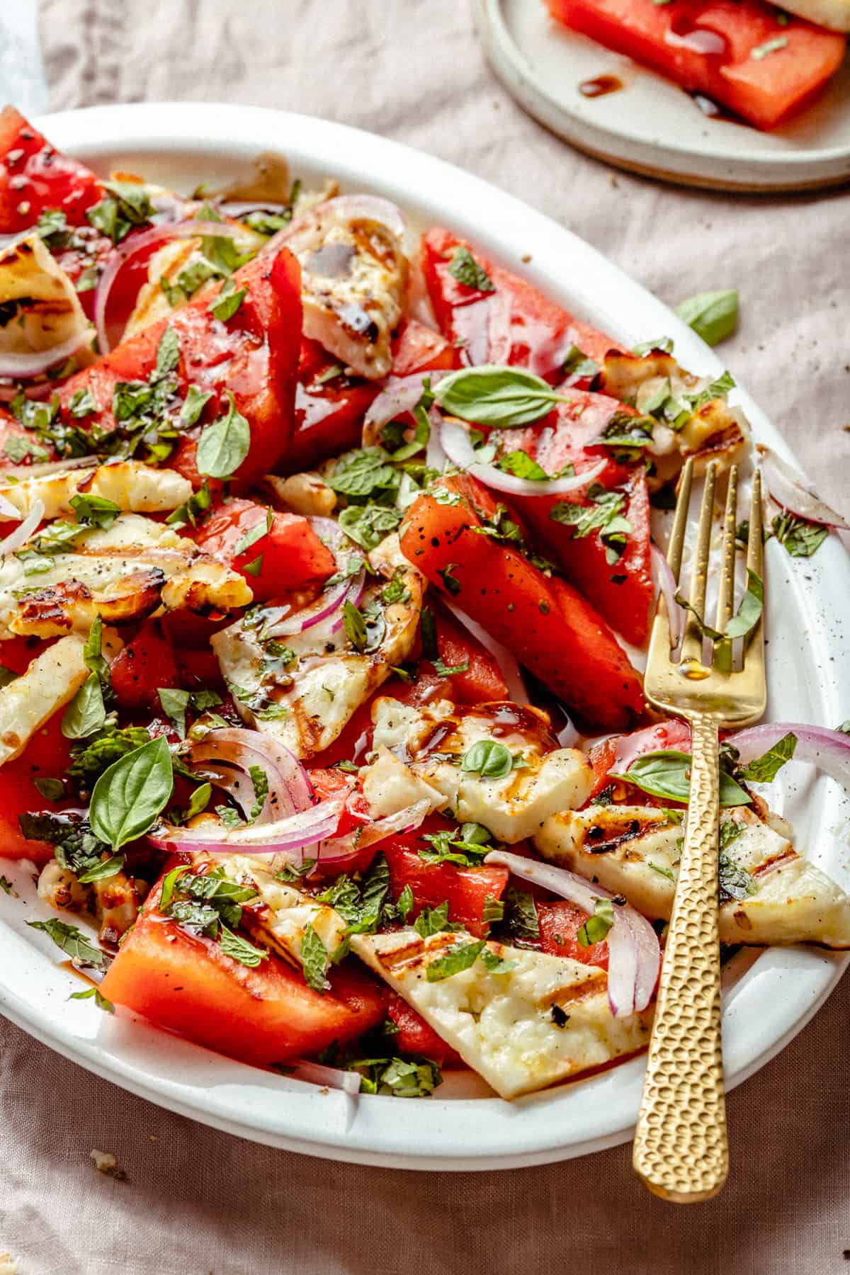 Close up showing the dressed watermelon basil salad with herbs, red onion slices and a golden fork to the side.