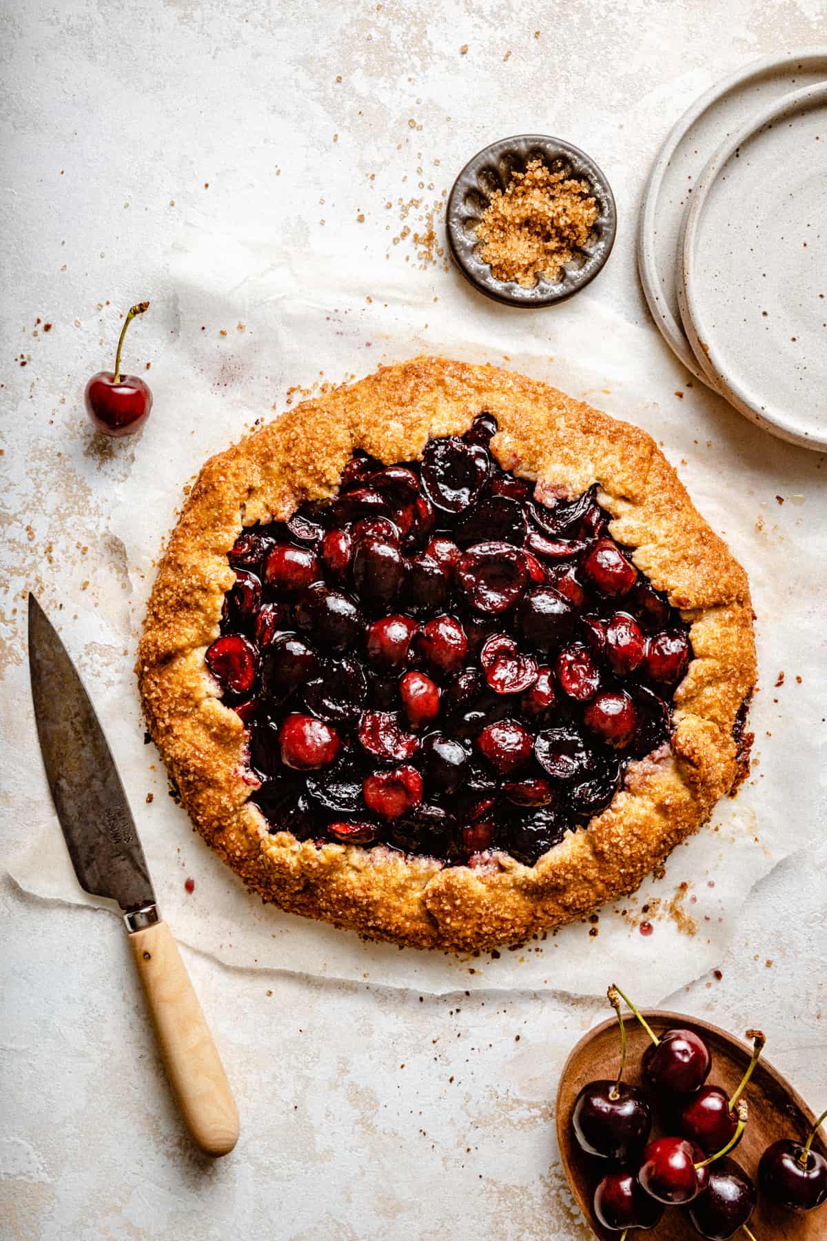 A galette filled with glistening red cherries on parchment paper with a knife to the side. 