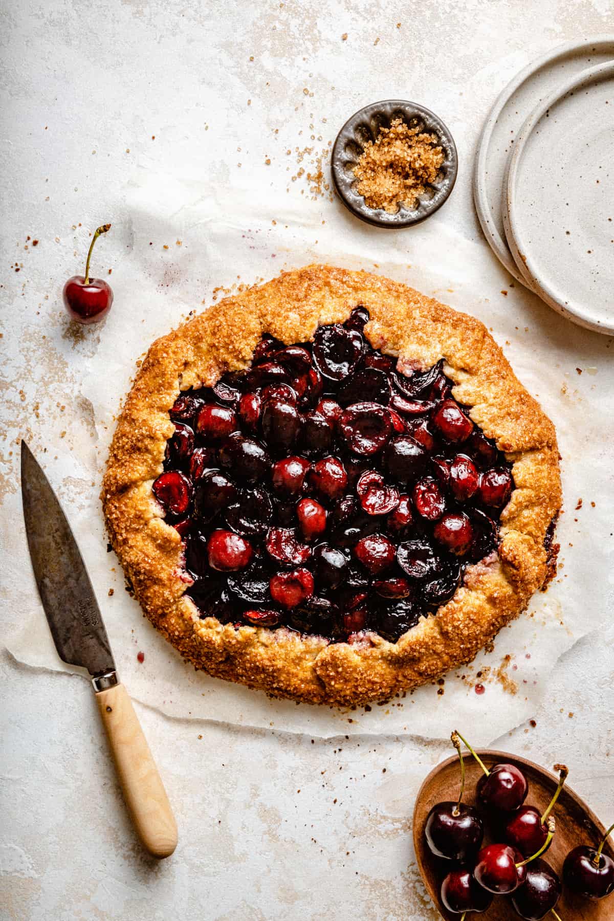 A galette filled with glistening red cherries on parchment paper with a knife to the side.