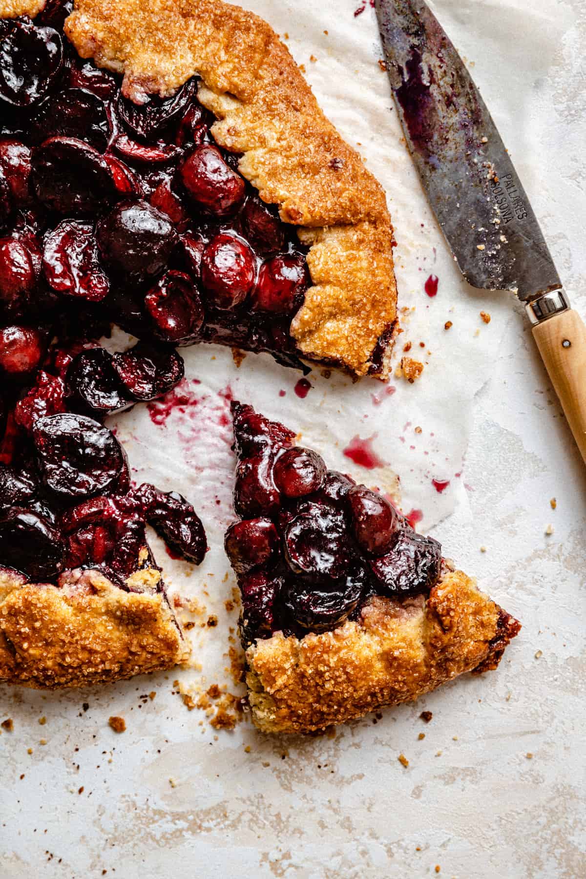 A close up of a slice taken from a cherry galette on parchment with a dirty knife to the side.