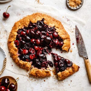 A cherry galette with two slices cut and a knife to the side with cherries around by.