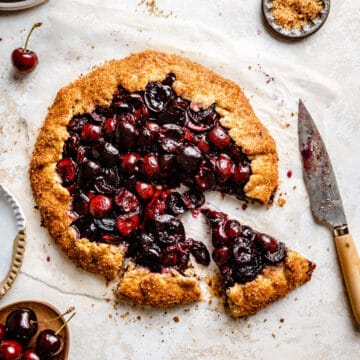 A cherry galette with two slices cut and a knife to the side with cherries around by.