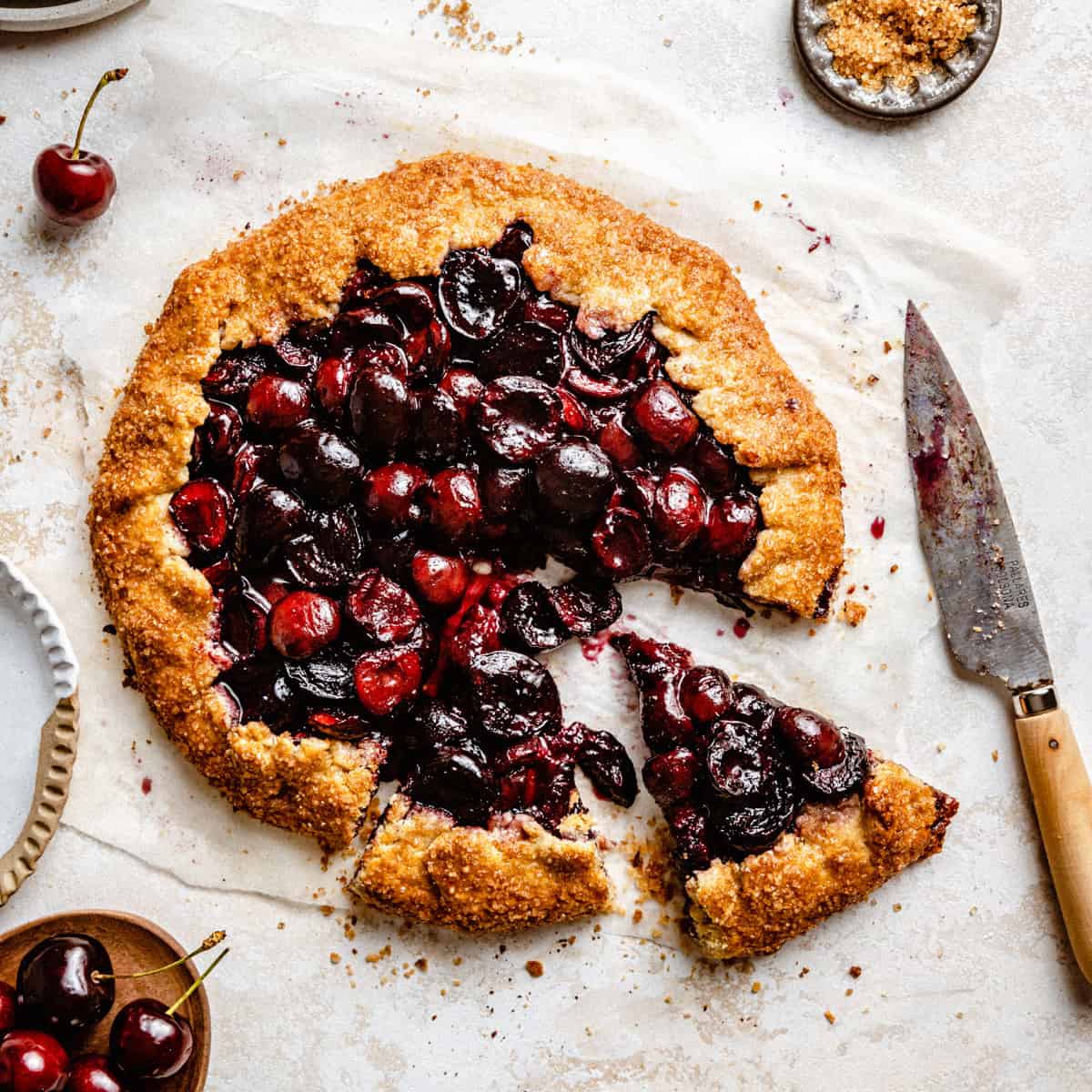 A cherry galette with two slices cut out on parchment with a knife next to it.