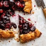 Cherry galette on a parchment background with a wedge cut out and a knife.