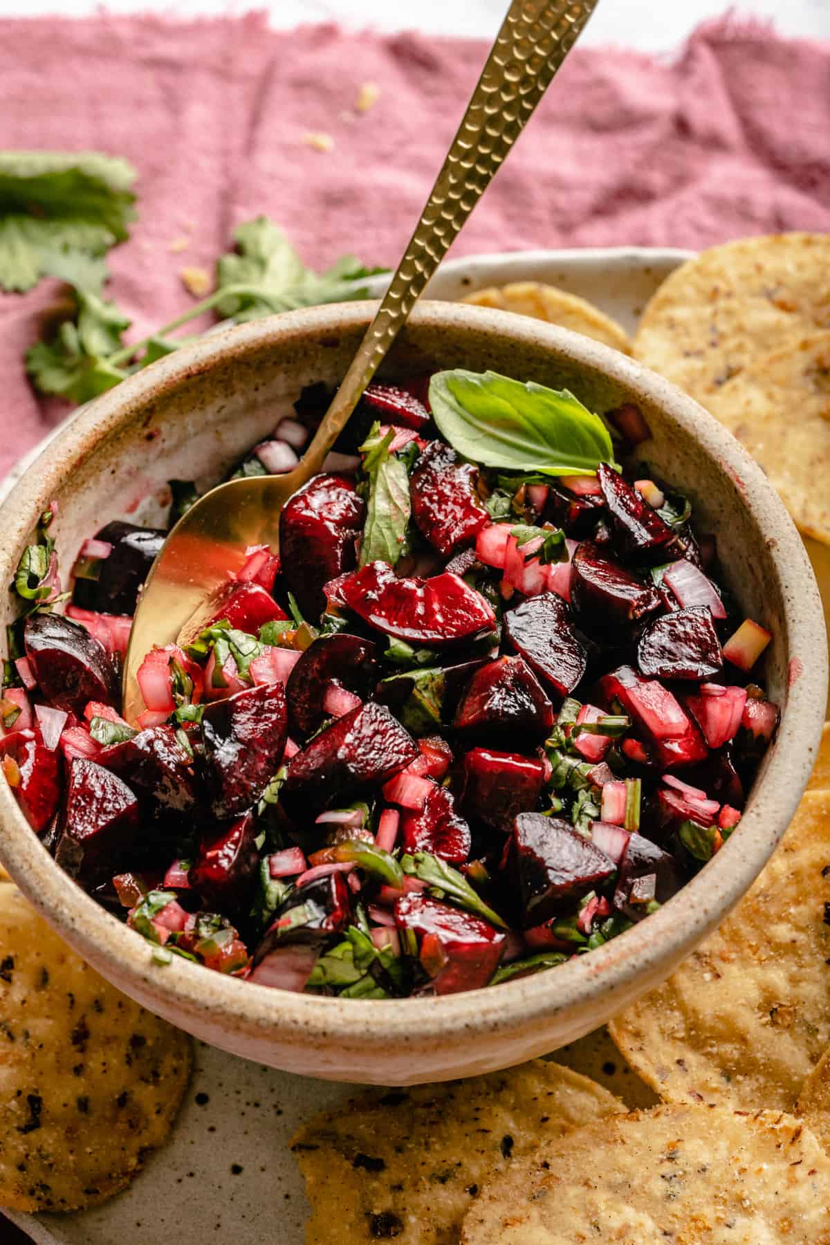 A close up of a bowl of cherry salsa with a golden spoon in an herbs on top.