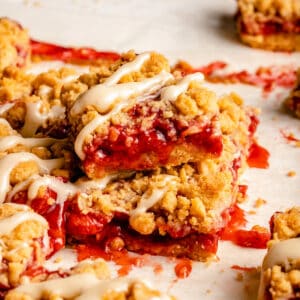 A stack of strawberry crumble bars with crumbs around and a creamy glaze on top.